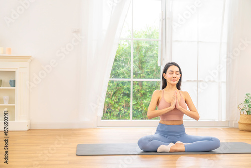 Happy Asian woman in sportswear practicing yoga wellness exercise Asana pose and meditation on yoga mat in bright room. Healthy woman enjoy sport training workout body stretching relaxation at home.