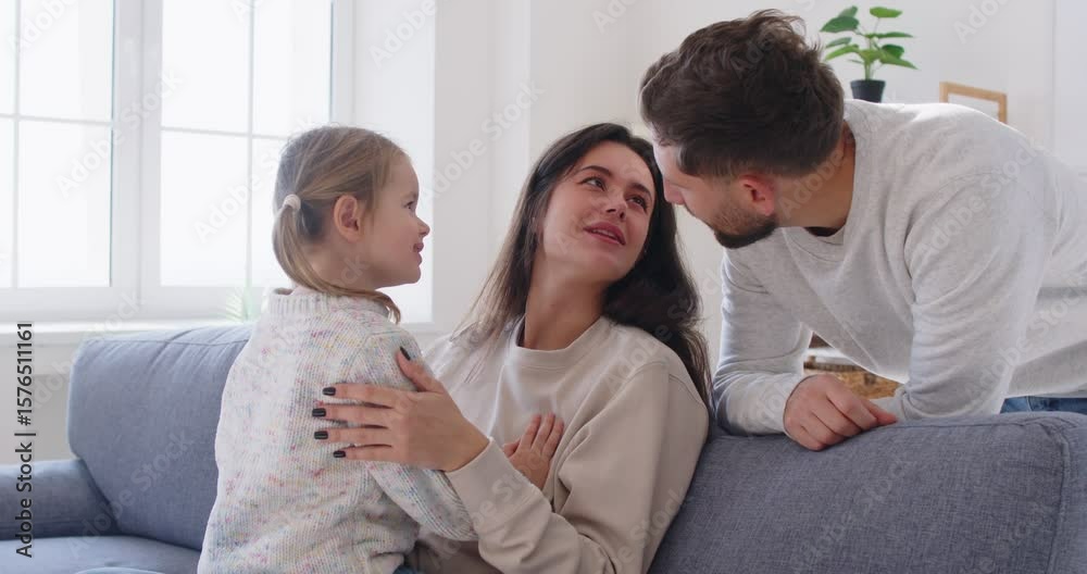 Smiling mother sitting on sofa, holding little preschool daughter on her knees while father standing behind them. Happy family spending quality time together at home, sharing tender and loving moment. Smiling mother sitting on sofa, holding little preschool daughter on her knees while father standing behind them. Happy family spending quality time together at home, sharing tender and loving moment.