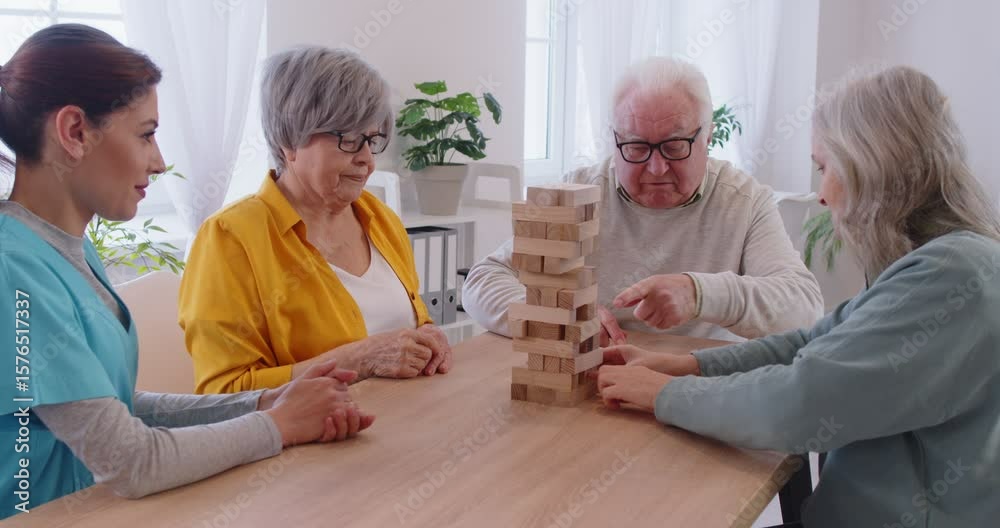 Group of elderly people sitting around table, enjoying board game and carefully removing blocks from wooden tower. Happy seniors playing jenga while young female caregiver watching them with smile.