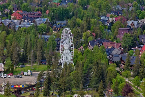 Fototapeta Naklejka Na Ścianę i Meble -  A white Ferris wheel rises above a green, tree-filled landscape. Colourful houses frame the scene, blending suburban charm with the whimsy of a quiet amusement setting in Zakopane - Lesser Poland - P
