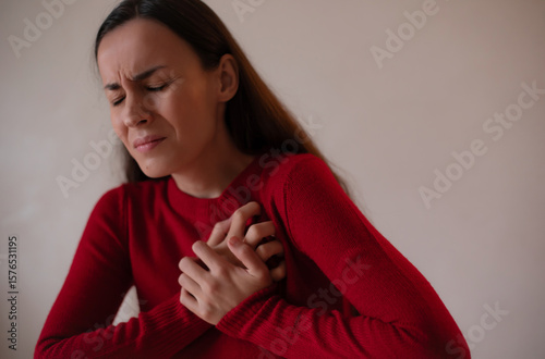 Young woman in a red sweater clutching her chest with eyes closed, expressing severe chest pain or heart attack symptoms, concept of heart health, stress, emergency.