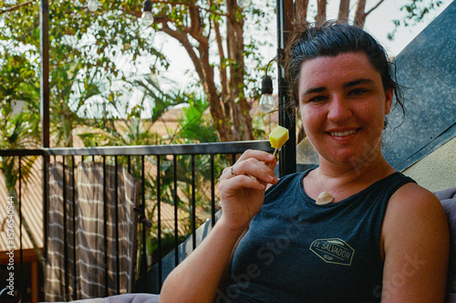 A cheerful woman seated outdoors enjoys a cubed pineapple snack on a patio surrounded by greenery, exuding relaxation. The casual setting features natural elements, creating a peaceful atmosphere.