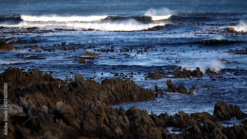 Scenic view of a dramatic rocky coastline with deep blue ocean waves crashing against the rugged shore. The lighting highlights textures of the rocks and creates a moody, cinematic atmosphere. Ideal f