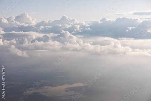Looking down on landscape of fields and farmland with clouds above. Serene vista from high above, showcasing clouds and land. Tranquil aerial scene of fluffy clouds over hazy landscape with water body