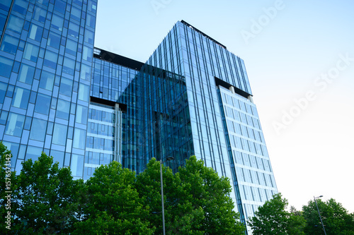 Bright modern skyscrapers with reflective glass facades rising above lush green trees under a clear sky — ideal for corporate power, finance, urban growth, and technology themes.