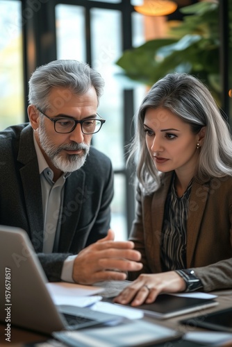 Team of diverse partners mature Latin business man and European business woman discussing project on laptop sitting at table in office. Two colleagues of professional business people working together.