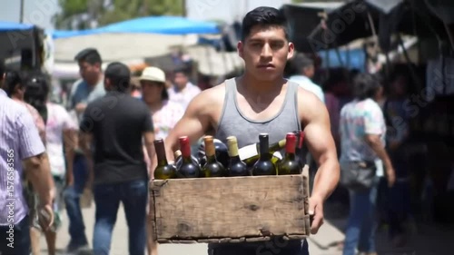 Man in tank top carries wooden crate filled with wine bottles through a bustling outdoor marketplace scene