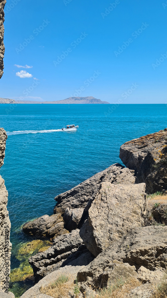 Obraz premium A scenic view of a turquoise sea with a small boat navigating through rocky cliffs under a clear blue sky. The landscape showcases natural beauty and tranquility.