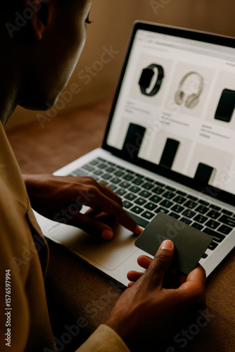 A person making an online purchase of electronic gadgets such as a smartwatch, headphones, and smartphones. The individual is using a laptop in a desk while holding a credit card