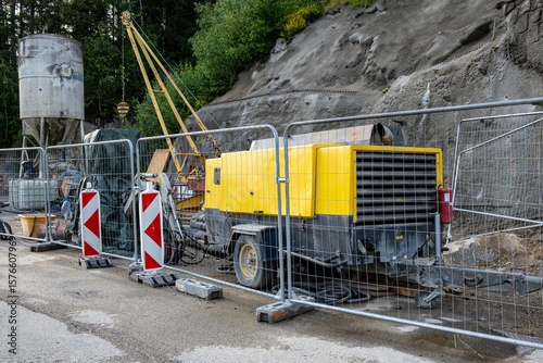 Mobile diesel air compressor and construction tools behind safety fencing. Road work zone with forested background. Industrial equipment on roadside.