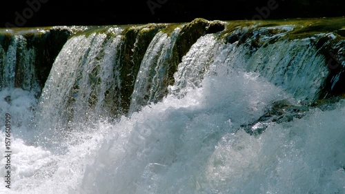 CLOSE UP, SLOW MOTION: Clear water cascades vigorously over a rocky edge covered with moss, making white foam and splashing drops. Raw power and mesmerizing natural beauty of a flowing, wild river.