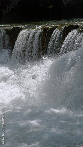 VERTICAL, CLOSE UP, SLOW MOTION: Small waterfall with dense jets of clear water that merge into a swirling pool below, creating abundant white foam and splashes. Raw energy of a free flowing river.