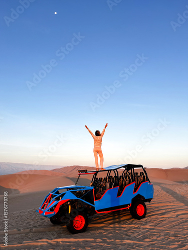 Woman standing on the roof top of buggy, that drives tourists around in desert area, while holding arm raised and watching beautiful arid landscape scenery Huacachina, Ica, Peru. Travel concept.