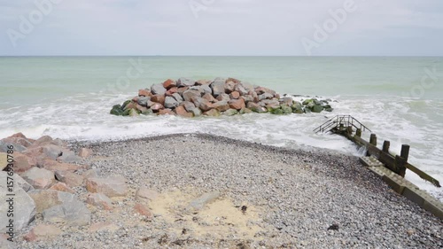 Wooden groynes and rocky breakwater on Cromer beach with waves washing over pebbles. Coastal defence structures protecting the Norfolk shoreline from erosion