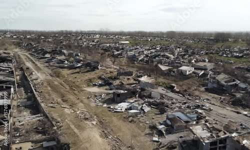 Drone footage flying over rows of collapsed houses, cracked roads and burnt walls, post-war scene, muted color grading