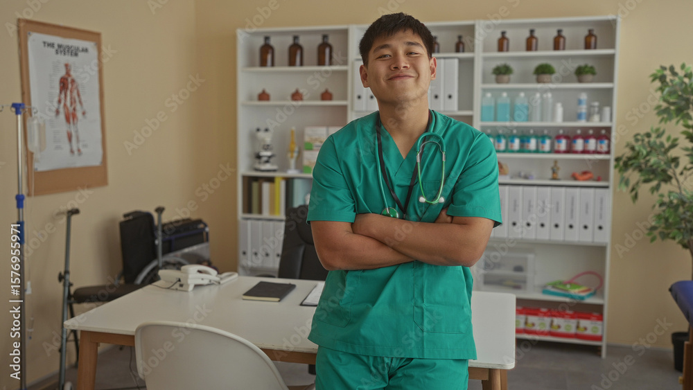 Obraz premium Young chinese doctor in green scrubs with arms crossed in a clinical room adorned with medical supplies, conveying a professional yet relaxed atmosphere.