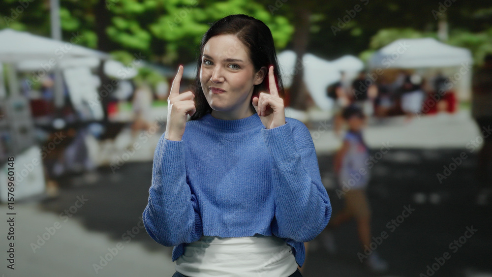 Woman outdoors in blue sweater points upward on a sunny day with market stalls in the background, expressing curiosity and focus in a vibrant street scene. Woman outdoors in blue sweater points upward on a sunny day with market stalls in the background, expressing curiosity and focus in a vibrant street scene.