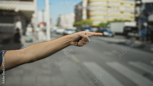 Man's hand pointing towards a bustling urban street with blurred city buildings and traffic, suggesting direction and focus amidst the vibrant outdoor setting.