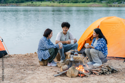 A group of friends sitting by the lake around a campfire, chatting and brewing coffee in front of an orange tent. Warm, relaxed outdoor camping atmosphere close to nature.