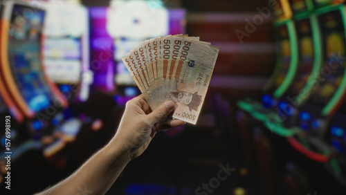 Man holding south korean won banknotes inside a casino with colorful slot machines in the background, highlighting the thrill of gambling and money handling.