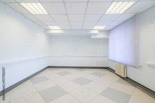 empty, brightly lit room with white walls, tiled floors, and vertical blinds on the window. The room has a drop ceiling and a radiator