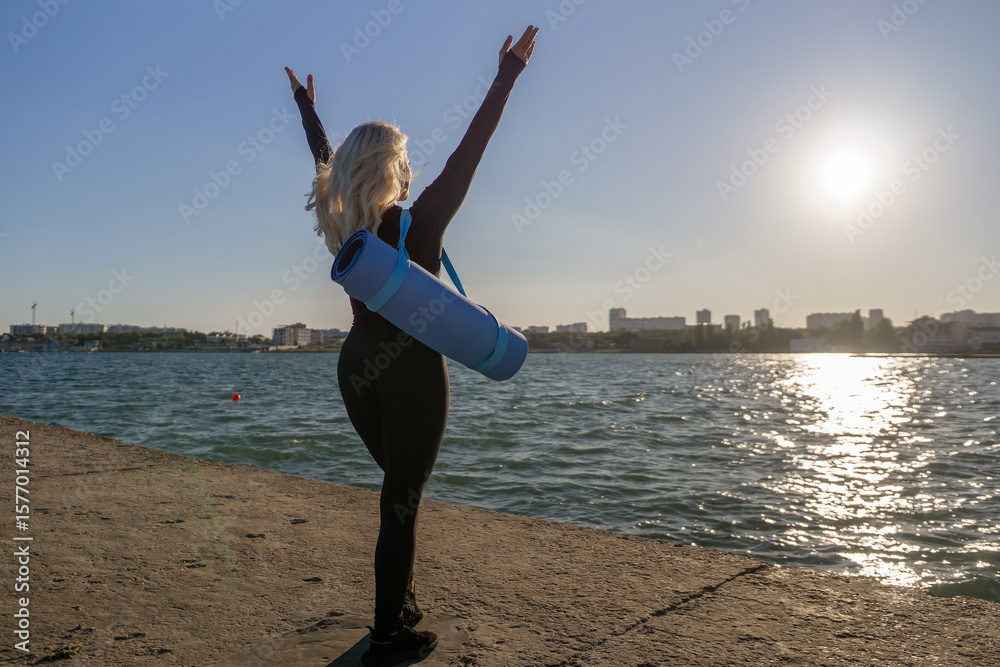 Obraz premium Woman Yoga Mat Beach Sunset: A woman in black sportswear raises her arms in celebration while standing on a beach with her yoga mat. The sun is setting behind her, casting a golden glow over the water