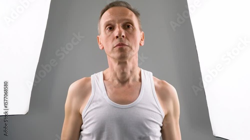 An elderly thin man in a white T-shirt does exercises. He lifts his shoulders up and down.