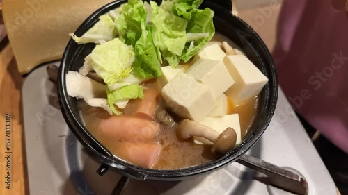 A close up view of a simmering hot pot filled with mushrooms, tofu, sausages, and fresh green leafy vegetables being added