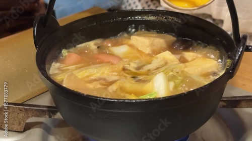 A close up shot shows a hand pouring a stream of beaten egg into a bubbling hot pot filled with various ingredients