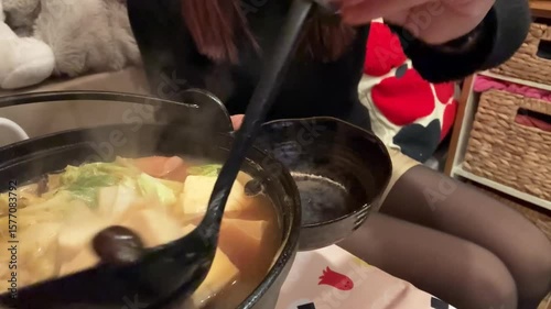 A person scoops various ingredients, including tofu, vegetables, and sausages, from a steaming hot pot into a smaller bowl using a large black ladle