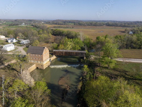Aerial view of the sunlit mill building casting shadows on the flowing river, with a covered bridge standing guard, Missouri, United States.