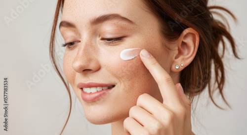 A woman with freckles applies cream to her cheek, showcasing a skincare routine for healthy, glowing skin