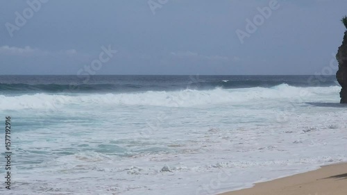 A serene tropical beach scene featuring the mesmerizing turquoise waters of the ocean gently meeting a sandy shore with foamy white waves. The vast blue sky is adorned with fluffy white clouds, sugges