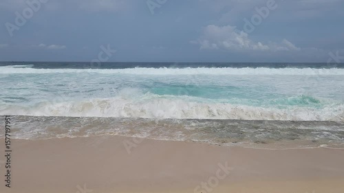A serene tropical beach scene featuring the mesmerizing turquoise waters of the ocean gently meeting a sandy shore with foamy white waves. The vast blue sky is adorned with fluffy white clouds, sugges