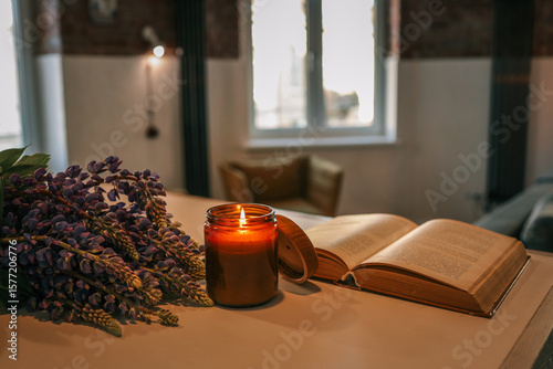 Lit candle, purple flowers, and an open vintage book on a light wooden table in a cozy loft-style room. Warm and peaceful atmosphere with natural light and rustic brick walls