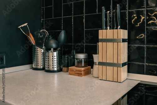 Modern kitchen countertop with black utensils in metal holders, wooden knife block, and spice jars against black tiled wall