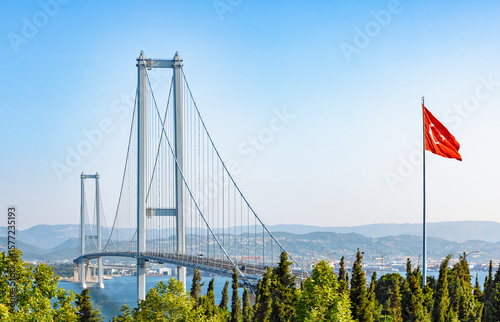 A majestic suspension bridge, the Osmangazi Bridge, spans a vast body of water, with vehicles crossing under a clear blue sky. A prominent Turkish flag proudly flies in the foreground.