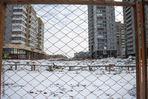 Snow covers a vacant construction site amid tall, modern buildings. A chain-link fence encloses the area, showcasing an incomplete urban project in a chilly environment.