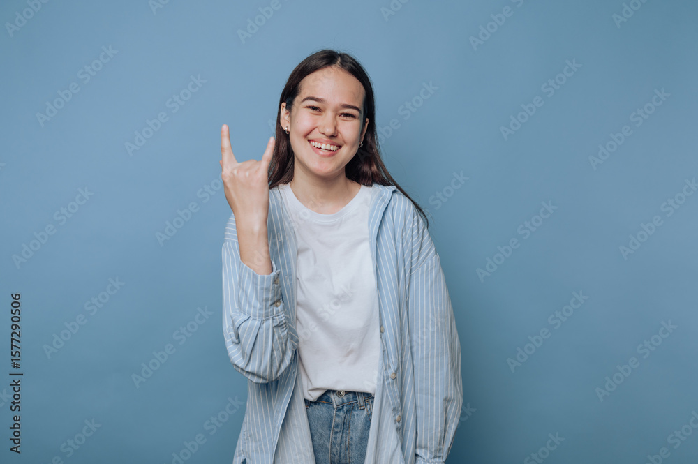 Fototapeta premium young woman joyfully poses with a rock on gesture, capturing a carefree spirit on light blue background