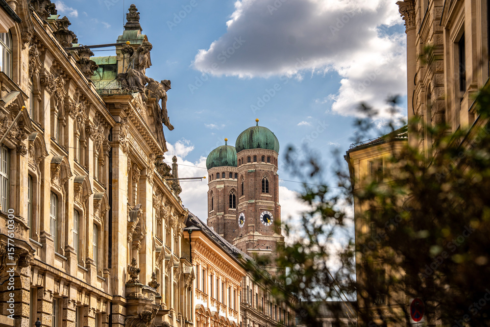 Fototapeta premium Iconic Cathedral Of Our Dear Lady In Munich: Majestic Twin Domes Rising Above Historic Buildings Under A Bright Blue Sky With Scenic Clouds Near Marienplatz