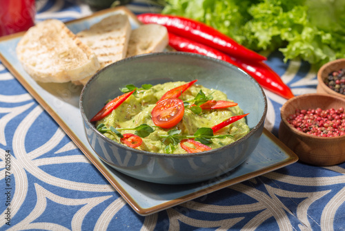 The photo shows a blue bowl filled with a creamy green dip, likely guacamole, garnished with red chili slices, a cherry tomato, and fresh microgreens. 