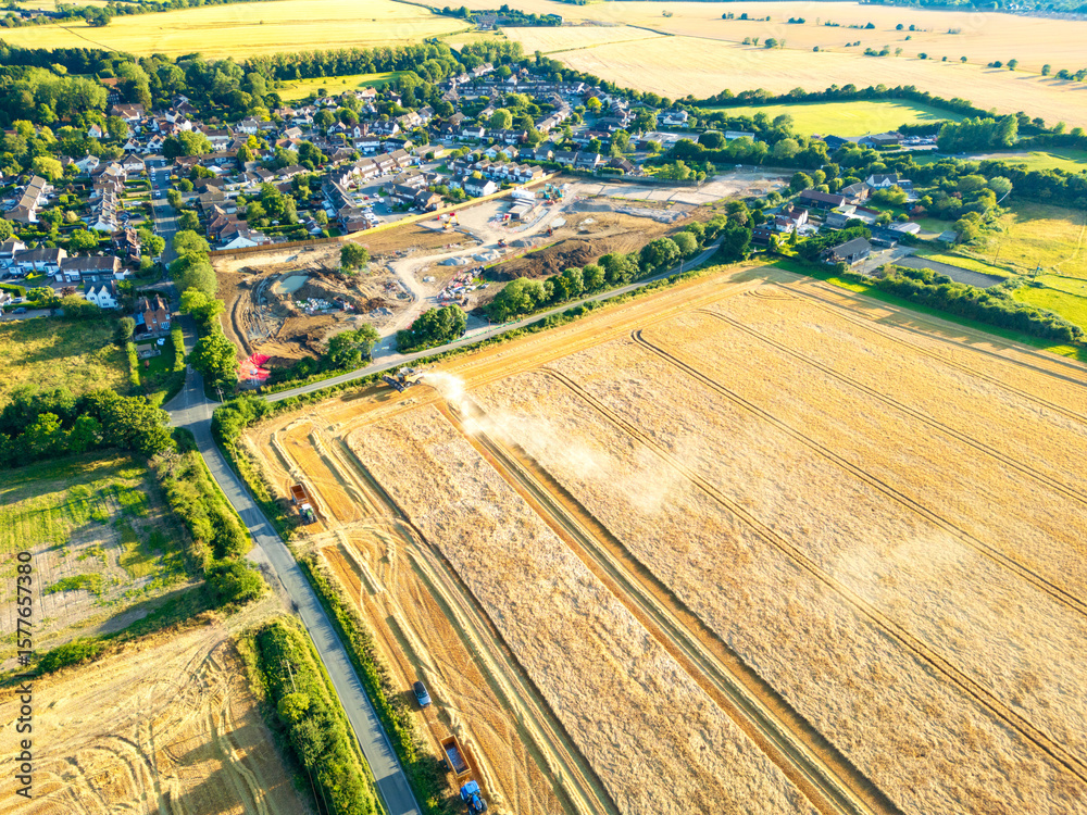 Fototapeta premium UAV view of a harvesting an arable field by a combine harvester in rural Essex, UK during late summer. A new housing development can be seen on the adjacent land.