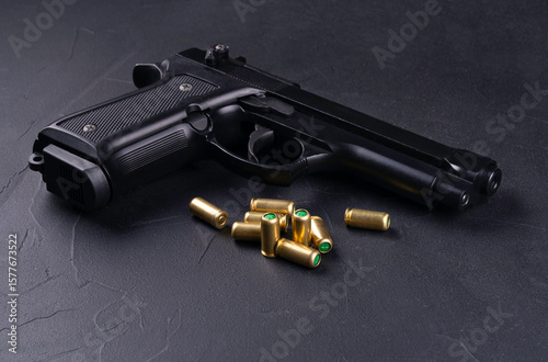 Black semi-automatic pistol with a textured grip lies on a dark surface next to several gold-colored bullets. The handgun is positioned at an angle, clearly displaying its barrel and trigger. This ima