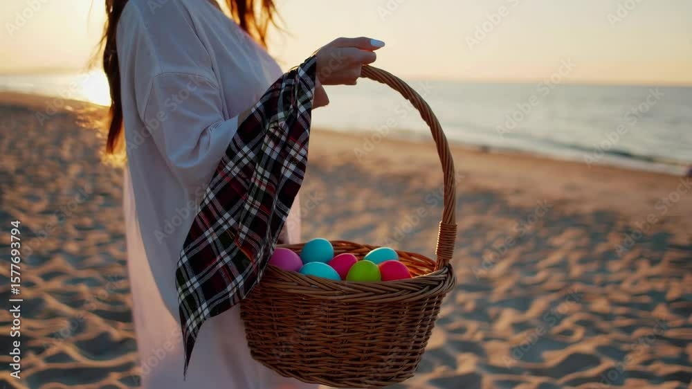 A woman carries a wicker basket with Easter Eggs, The setting sun and the wind blows the plaid on it. Sea sand and waves in the background