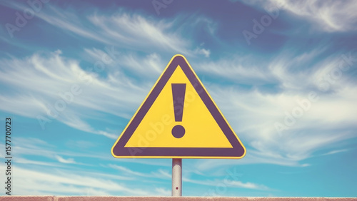 Warning yellow triangular sign bearing exclamation mark signaling potential hazard, positioned against overcast sky background