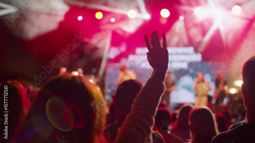 Person raises hand at outdoor concert or worship event, with colorful red stage lighting and blurred background. Ideal for music, celebration, church or festival atmosphere footage. 4K Backside shot