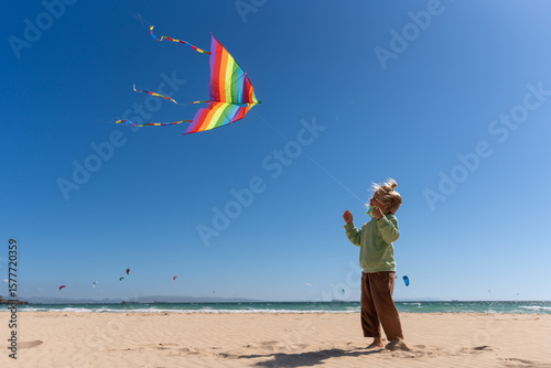 Fotografía Smiling young boy flying a colorful rainbow kite on a sunny sandy beach with blu