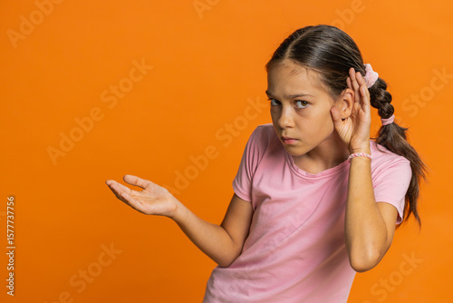I cant hear you. What. Child girl kid trying hear you, looking confused and frowning, keeping arm near ear for louder voice asking to repeat to hear information deafness. Children on orange background