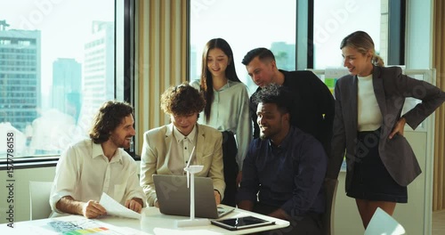 Diverse office team clapping and smiling around a laptop, celebrating success in a bright modern workspace with city view and creative planning materials.
