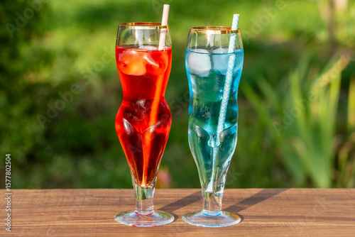 Two artistic glasses shaped like a nude woman and man filled with red and blue alcoholic cocktails on table with lush green garden background, closeup. Playful summer vibe for creative drink concepts